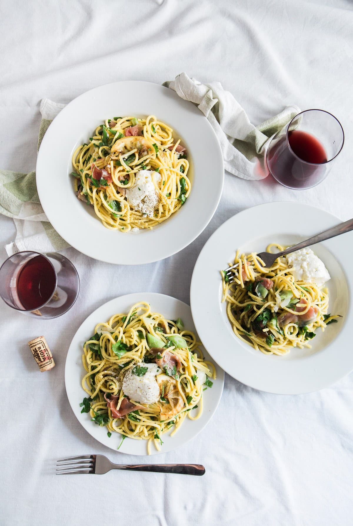 Four plates of pasta and glasses of red wine on a white tablecloth, from above