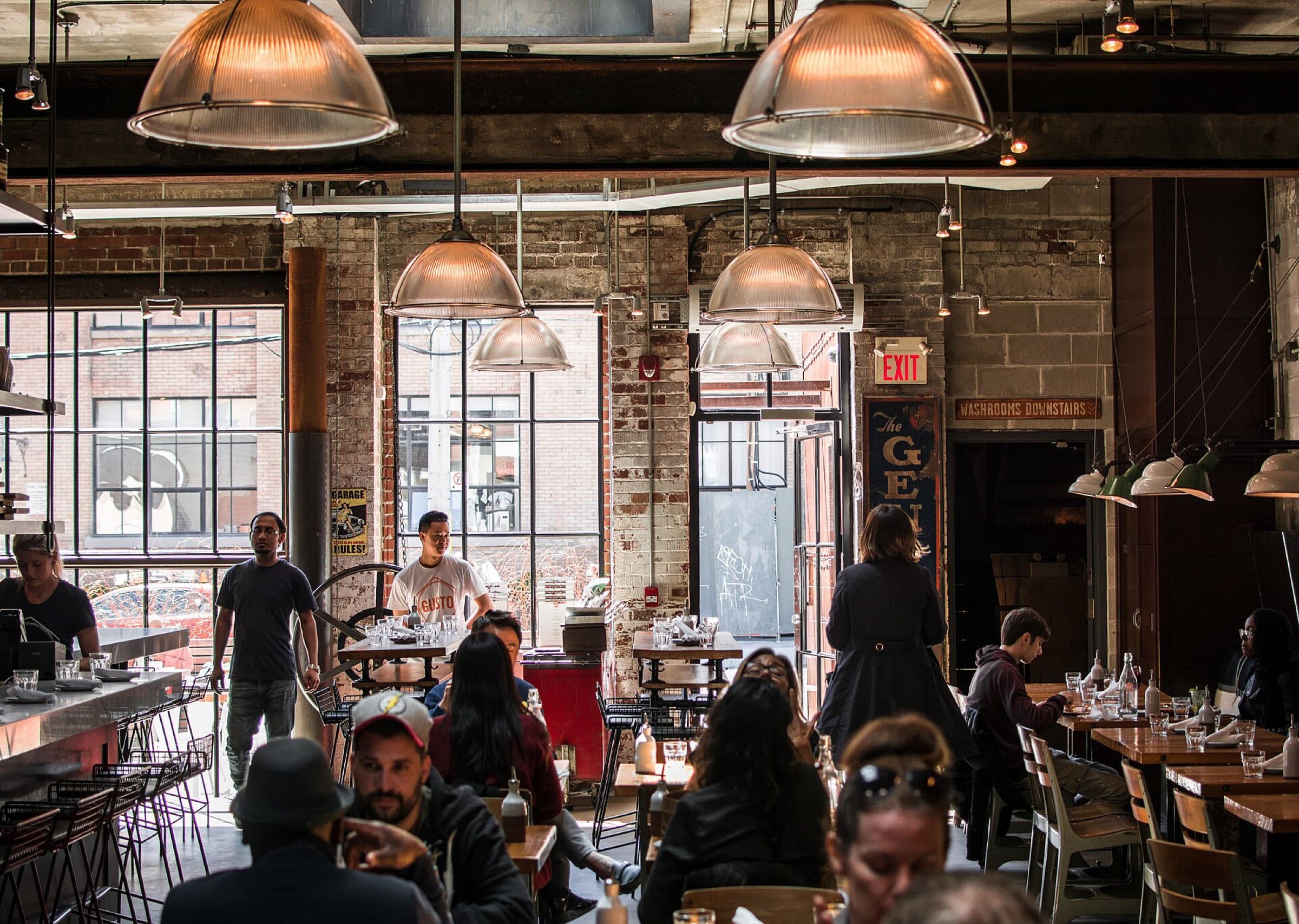 A busy restaurant with tall windows, brick walls and hanging lamps
