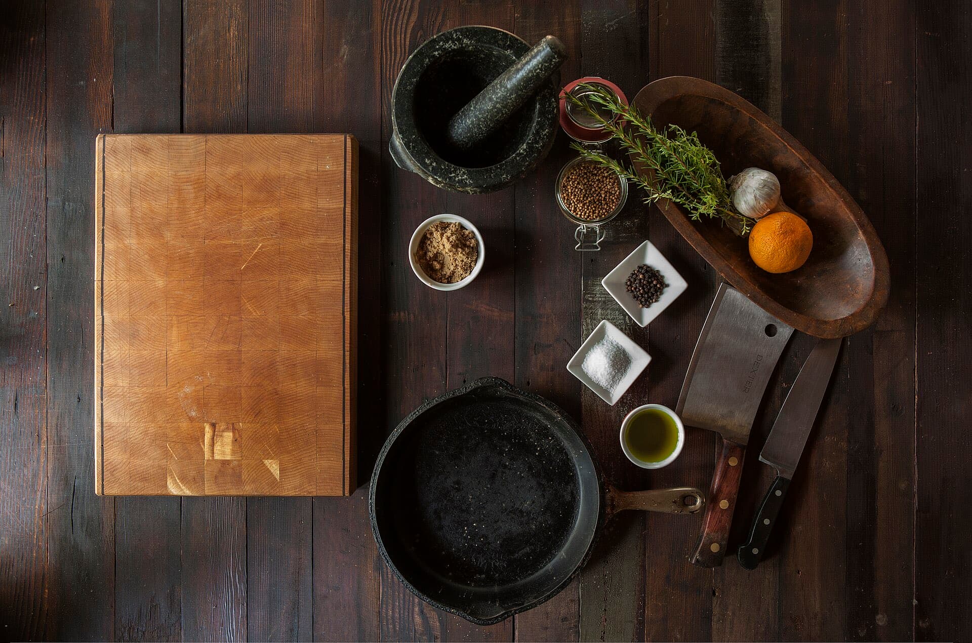 A chef's station from above: cutting board, mortar, herbs, spices and cast iron